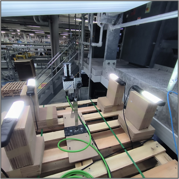 Câmera industrial montada em estrutura de paletes junto à linha da rotativa, cercada por quatro luminárias de LED apontadas para o ponto de captura